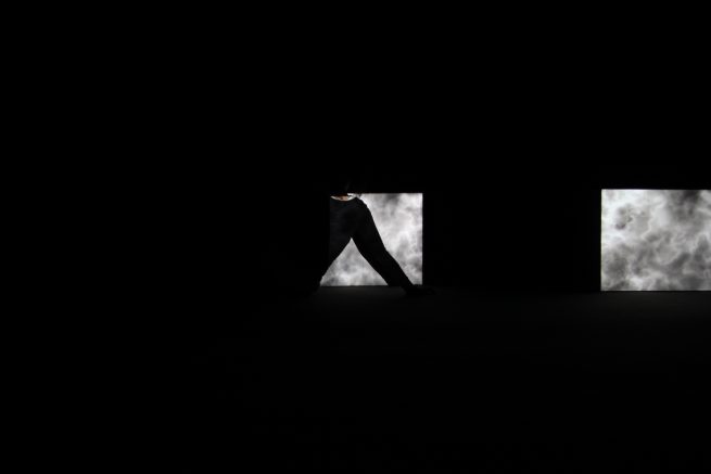 from the work silence. The image shows the performer standing in front of the installation. The installation was two projected square clouds and a projected noise on the background